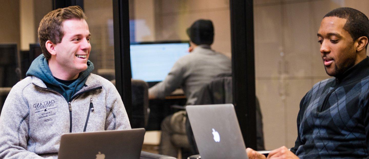 Two men seated with Mac laptops open in front of them